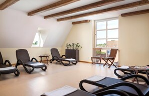 Ein Zimmer mit geneigter Decke, Holzbalken und großen Fenstern. Mehrere Lounge-Sessel und ein kleiner Tisch mit einer Pflanze sind vorhanden.