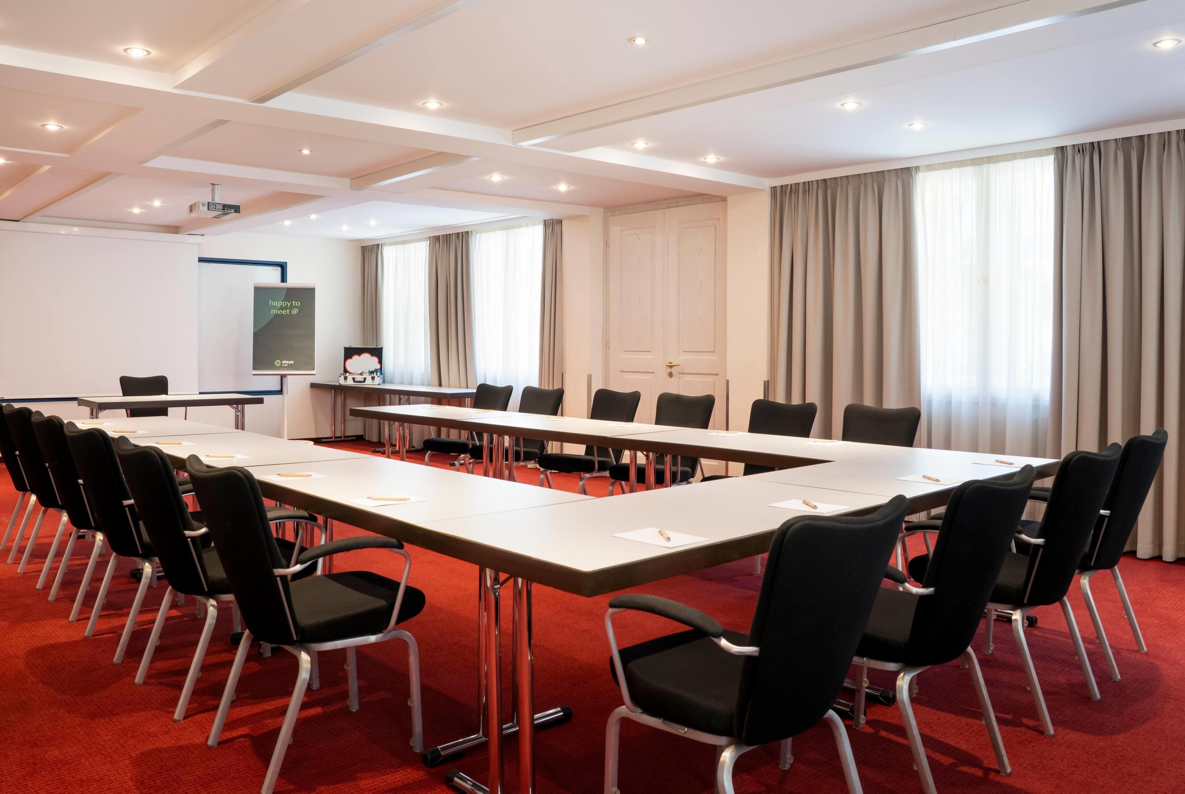 Ein Konferenzraum mit langem Tisch, schwarzen Stühlen, einer Whiteboard-Tafel und einer Projektionsleinwand.