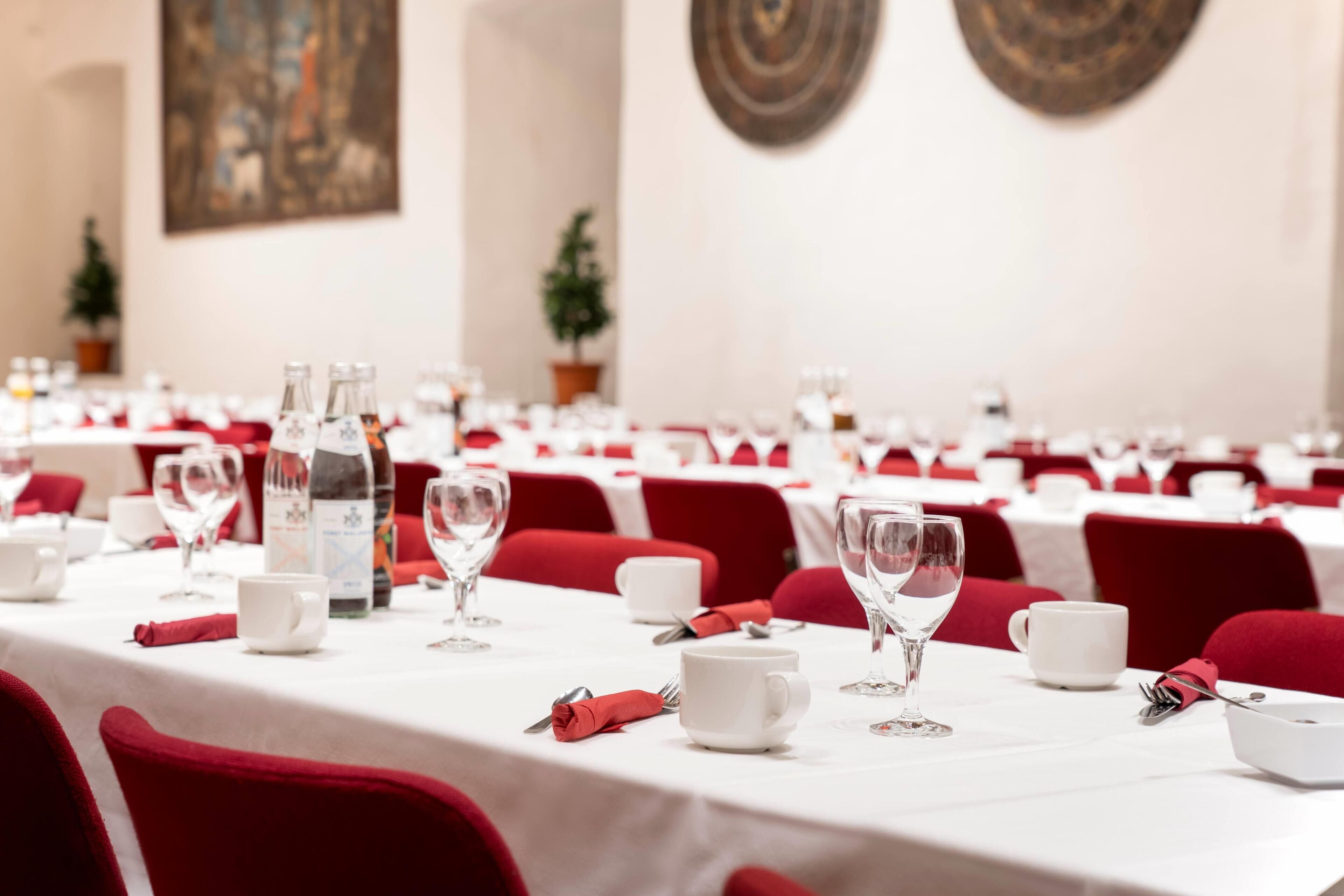 A banquet hall with tables set for dining, each with white tablecloths, red chairs, and clear glassware.