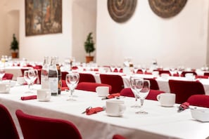 Een feestzaal met gedekte tafels, elk met witte tafellakens, rode stoelen en glazen.'