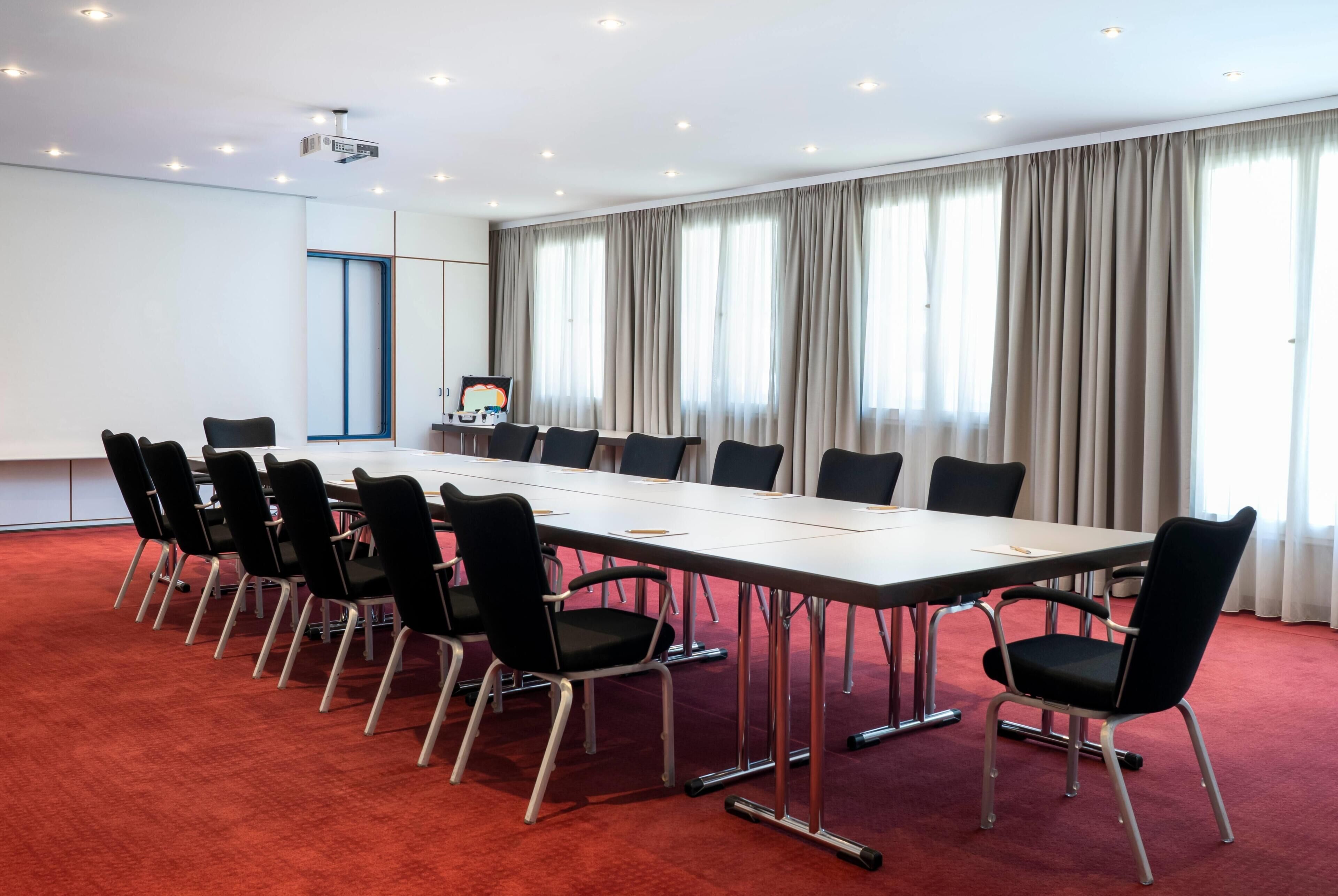 A conference room with a long table, black chairs, a projector, and a large window with curtains.