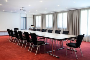 Een vergaderzaal met een lange tafel, zwarte stoelen, een projecteerder en een groot raam met gordijnen.