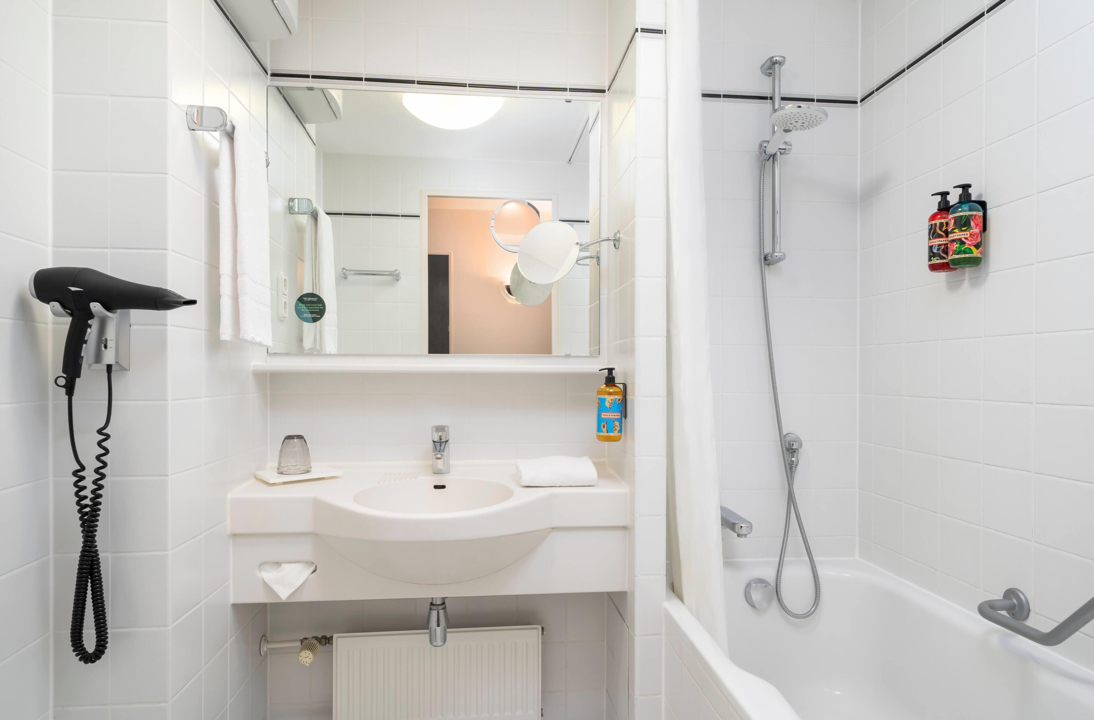 A bathroom with a white sink, a mirror, a shower, and a toilet paper dispenser.