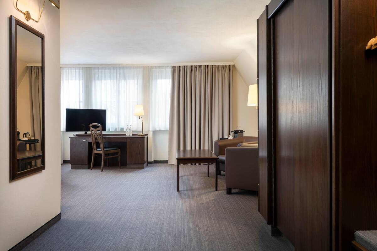 Habitación de hotel con escritorio, silla, televisión y un amplio ventanal con cortinas.