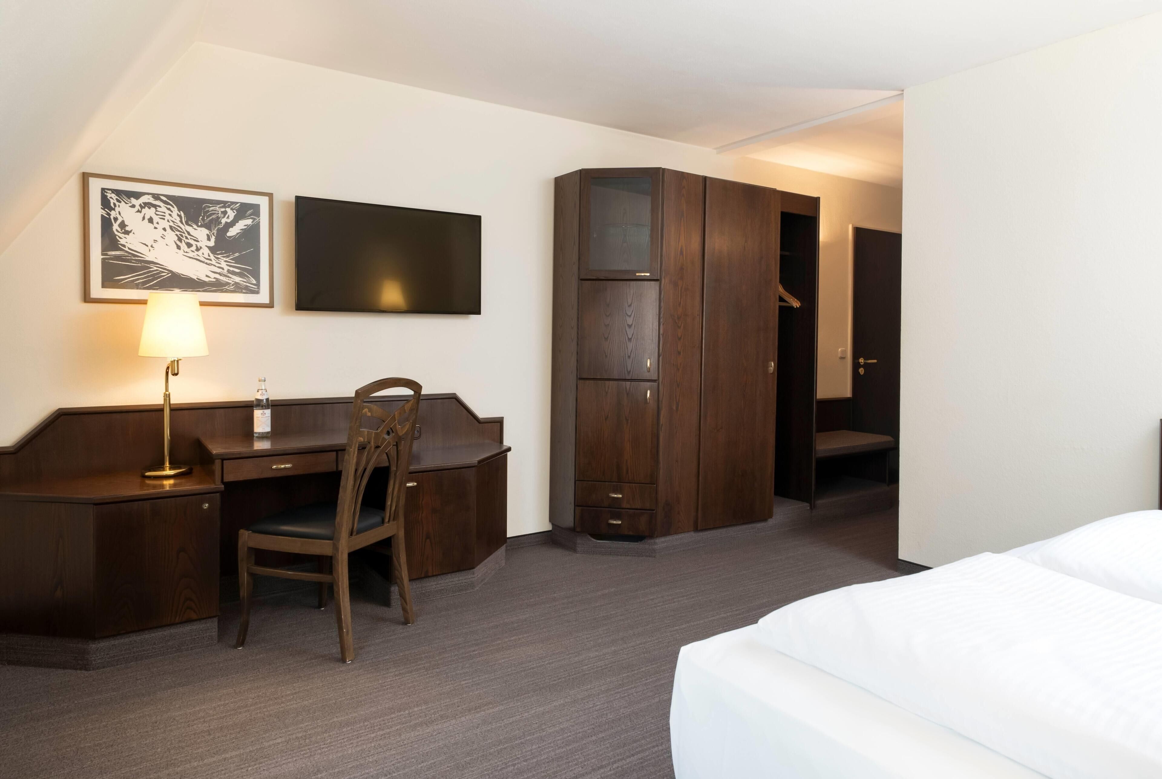 A hotel room with a bed, a desk with a chair, a television, and a wardrobe.