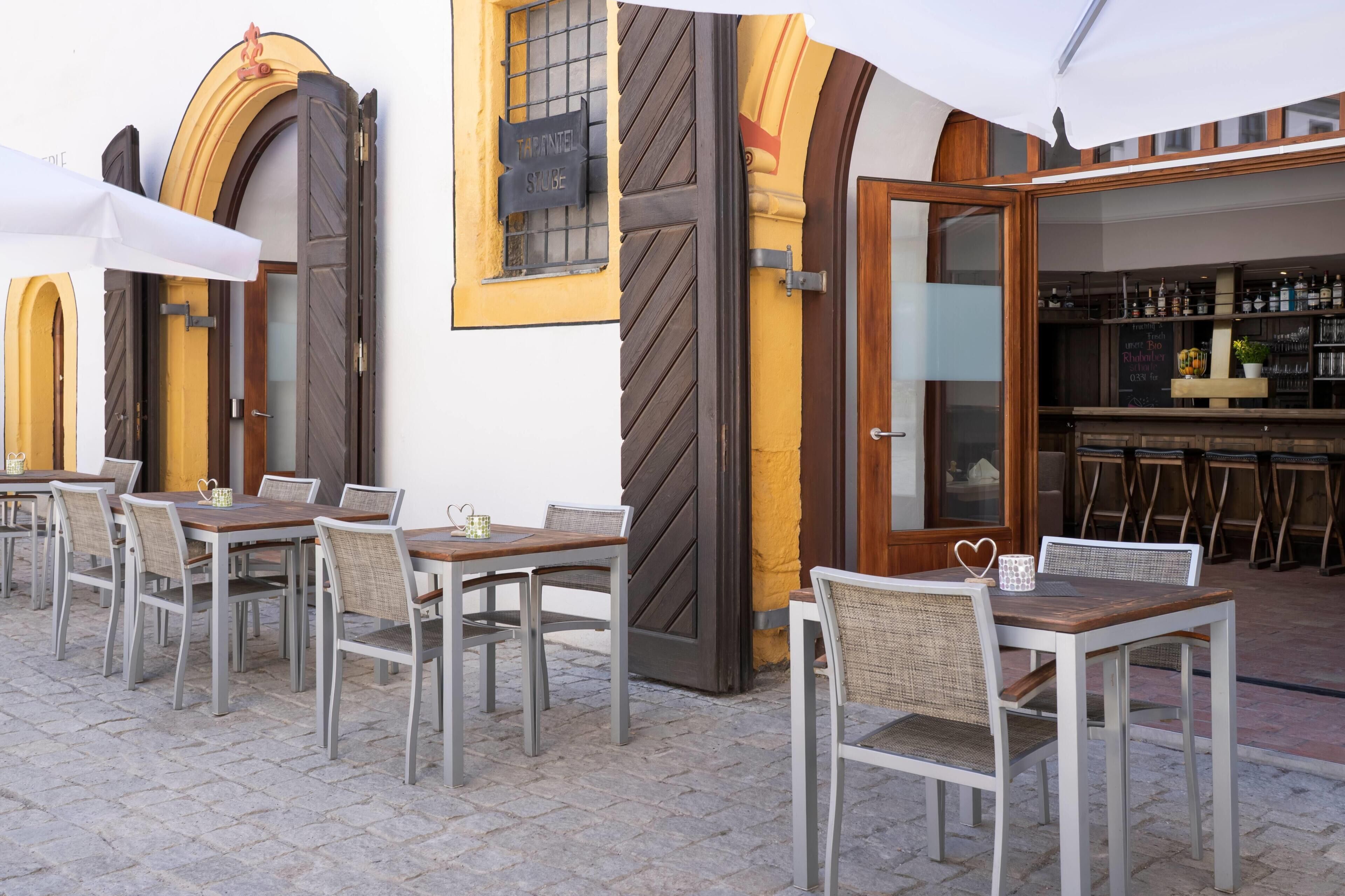 An outdoor cafe with tables and chairs, a bar area with bottles, and a yellow and brown facade.