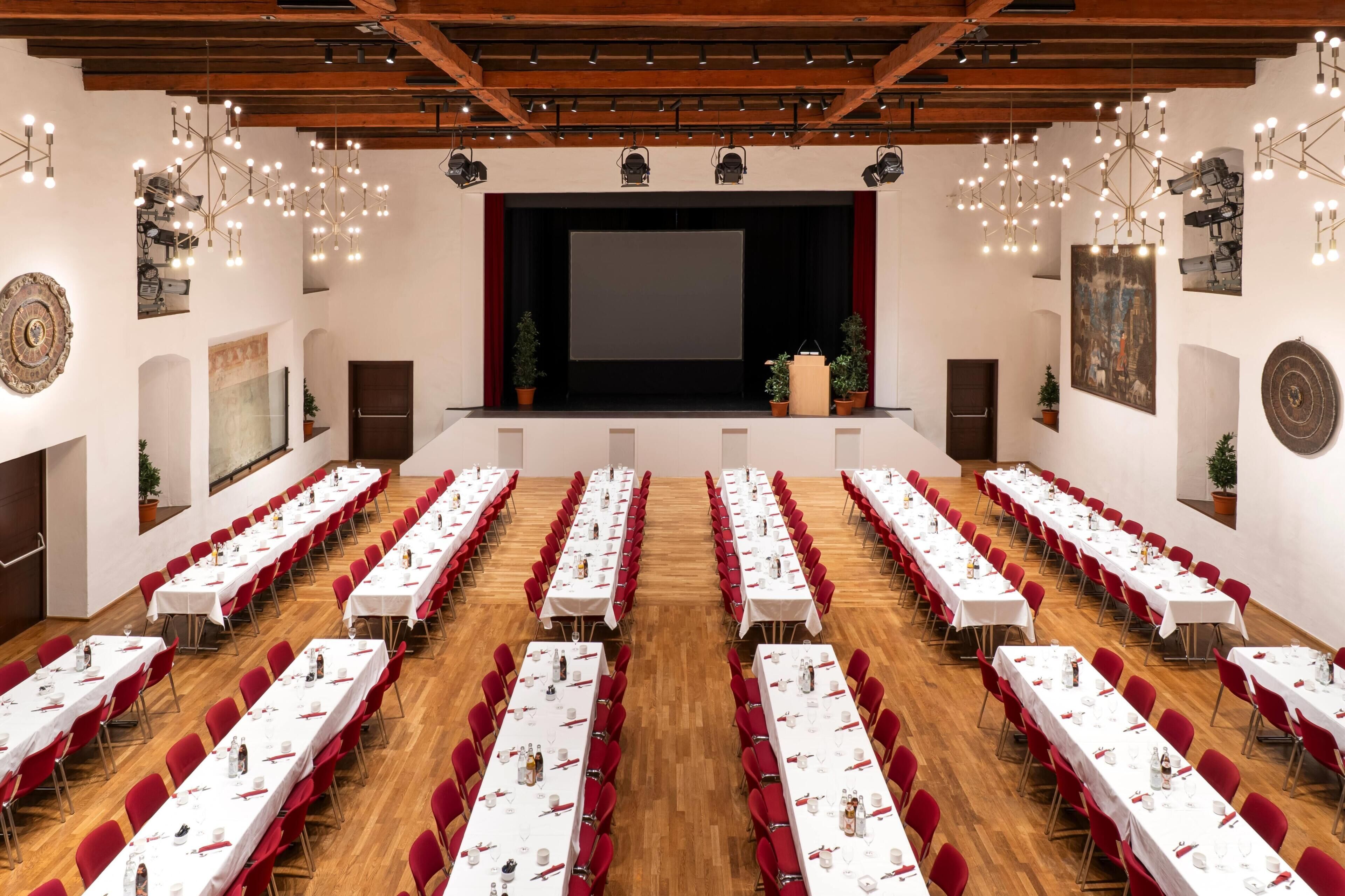 A conference room with multiple tables set for dining, a stage with a screen, and a wooden ceiling.