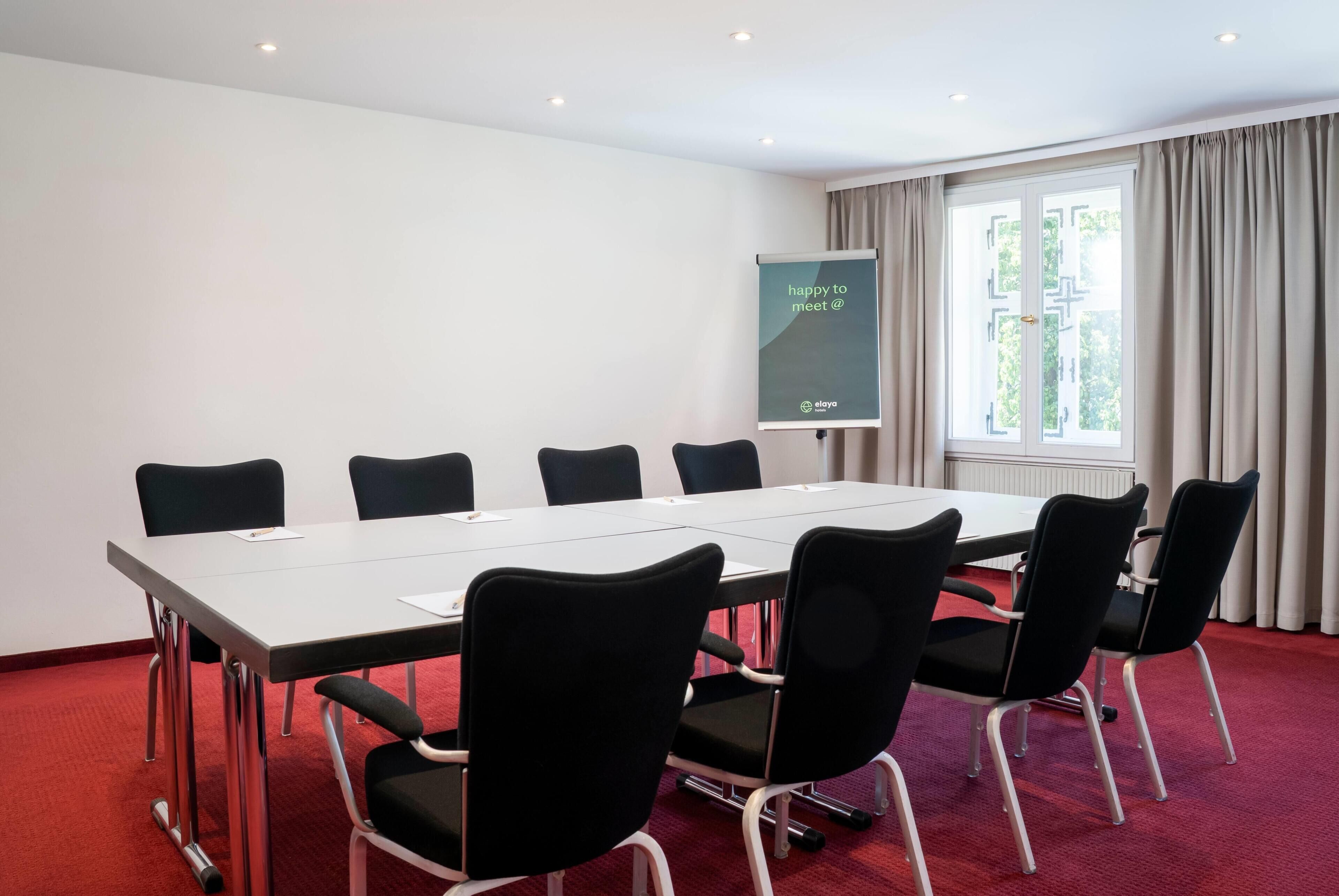 Un salón de conferencias con una larga mesa y seis sillas, una pantalla de proyector y un ventanal con cortinas.