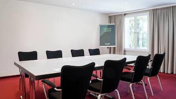 Un salón de conferencias con una larga mesa y seis sillas, una pantalla de proyector y un ventanal con cortinas.