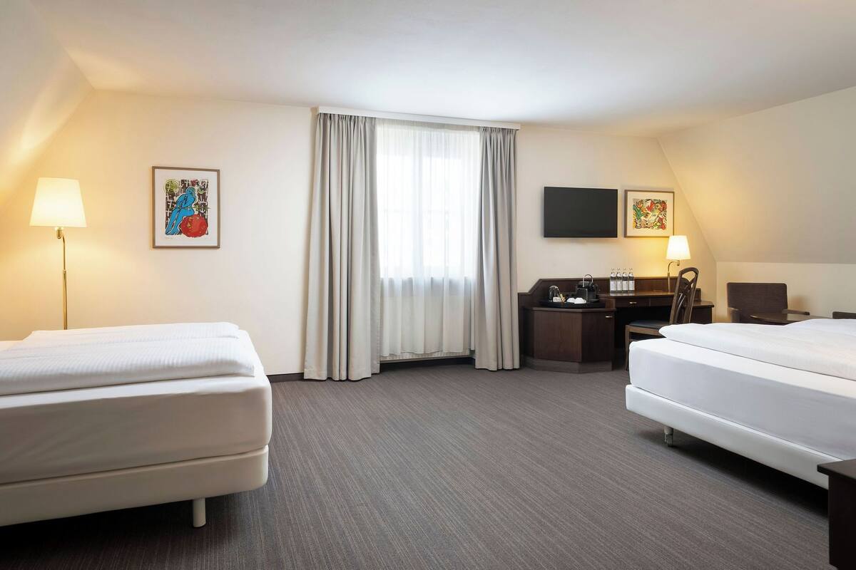 Habitación de hotel con dos camas, un escritorio, una silla, un televisor de pantalla plana y un ventanal con cortinas.