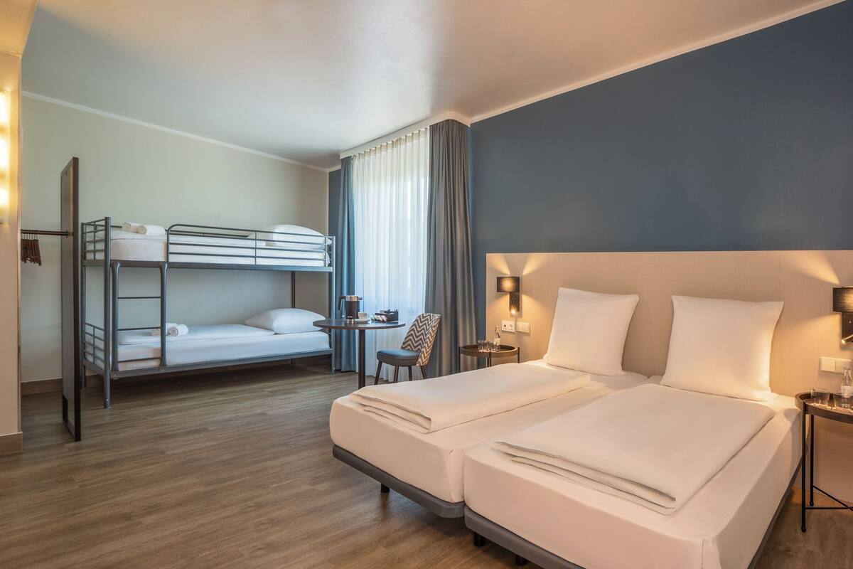 Habitación de hotel con literas, una cama doble, una mesita pequeña con una silla y una mesita de noche con lámpara.