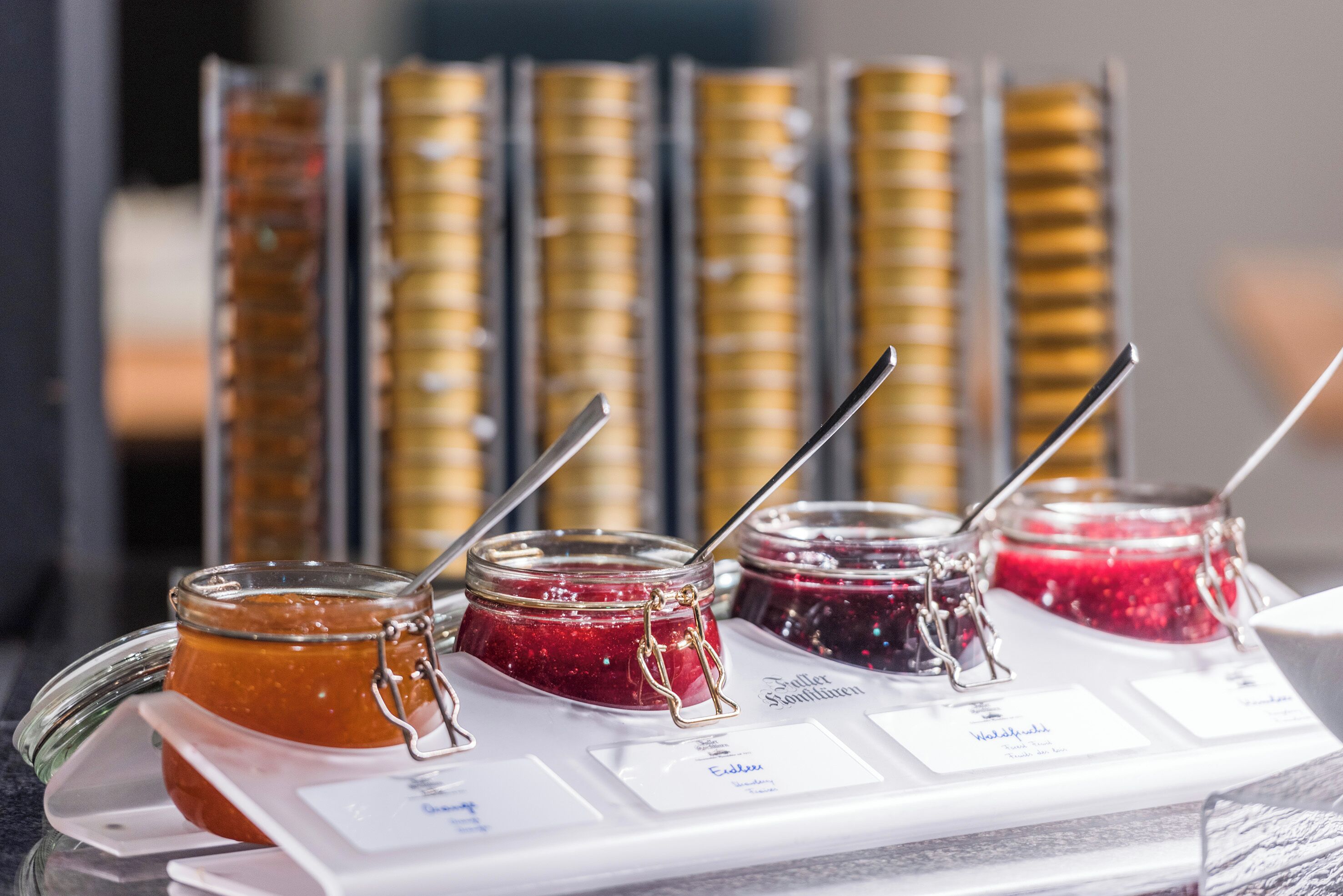 A display of various jams with labels, some jars with metal clasps and spoons, and a background of stacked jars.