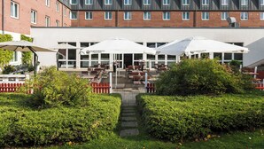Ein Gebäude mit einem Café-Bereich, der Außenplätze und Sonnenschirme bietet, umzäunt mit einem roten Ziergartenzaun und gesäumt von gepflegten Hecken.