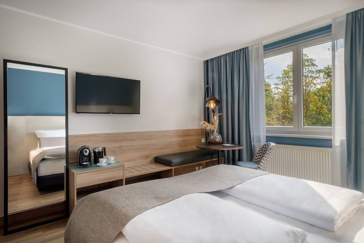 Una habitación de hotel moderna con una cama grande, una mesita de noche con lámpara, televisión y vistas a árboles a través de la ventana.