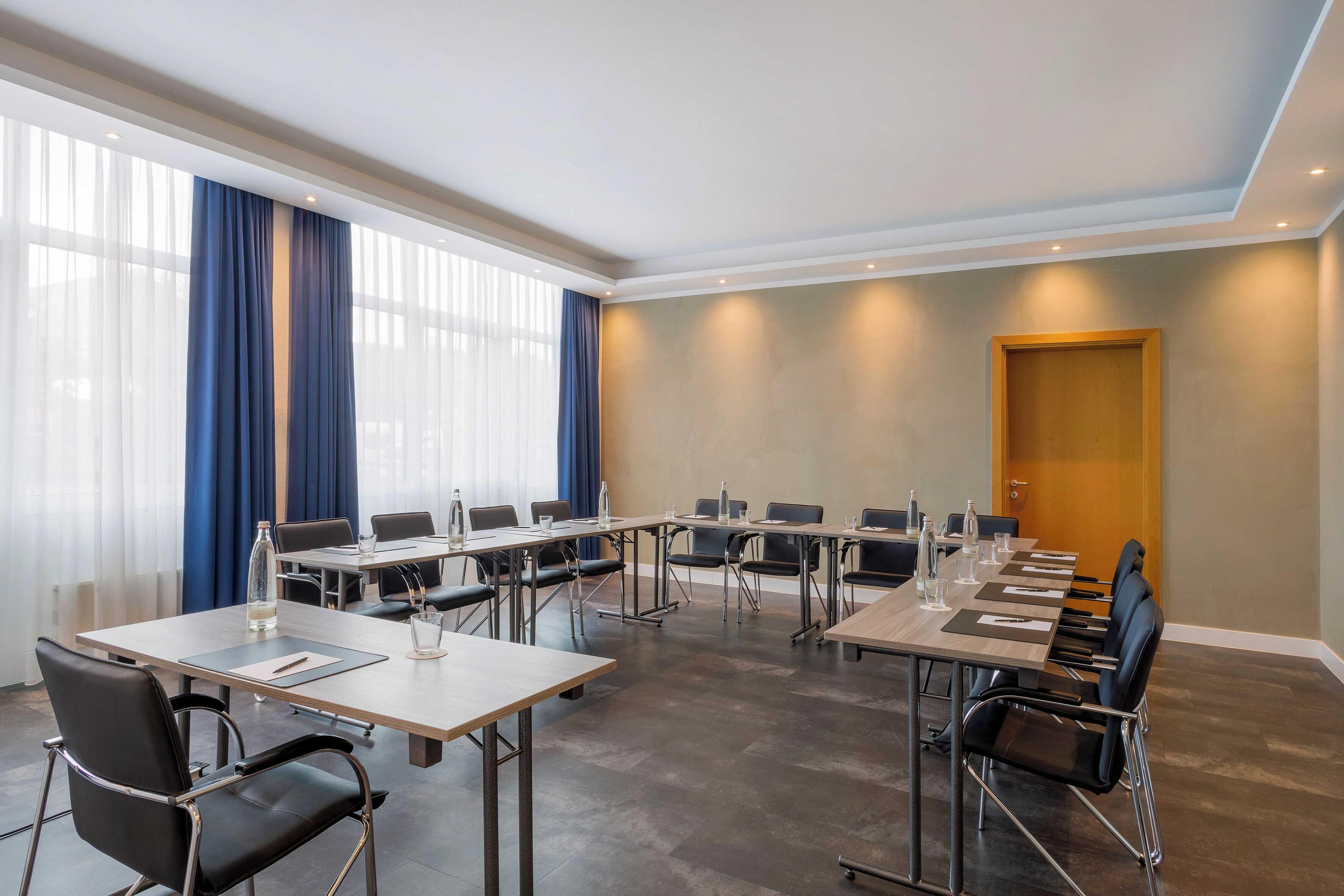 Ein Konferenzraum mit mehreren Tischen und Stühlen, großen Fenstern mit Vorhängen und einer Tür.