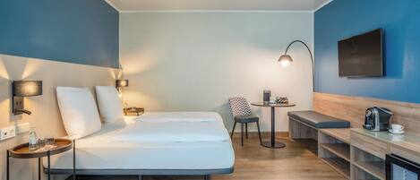 A modern hotel room with a large bed, bedside tables, a small seating area, a coffee machine, and a flat-screen TV.