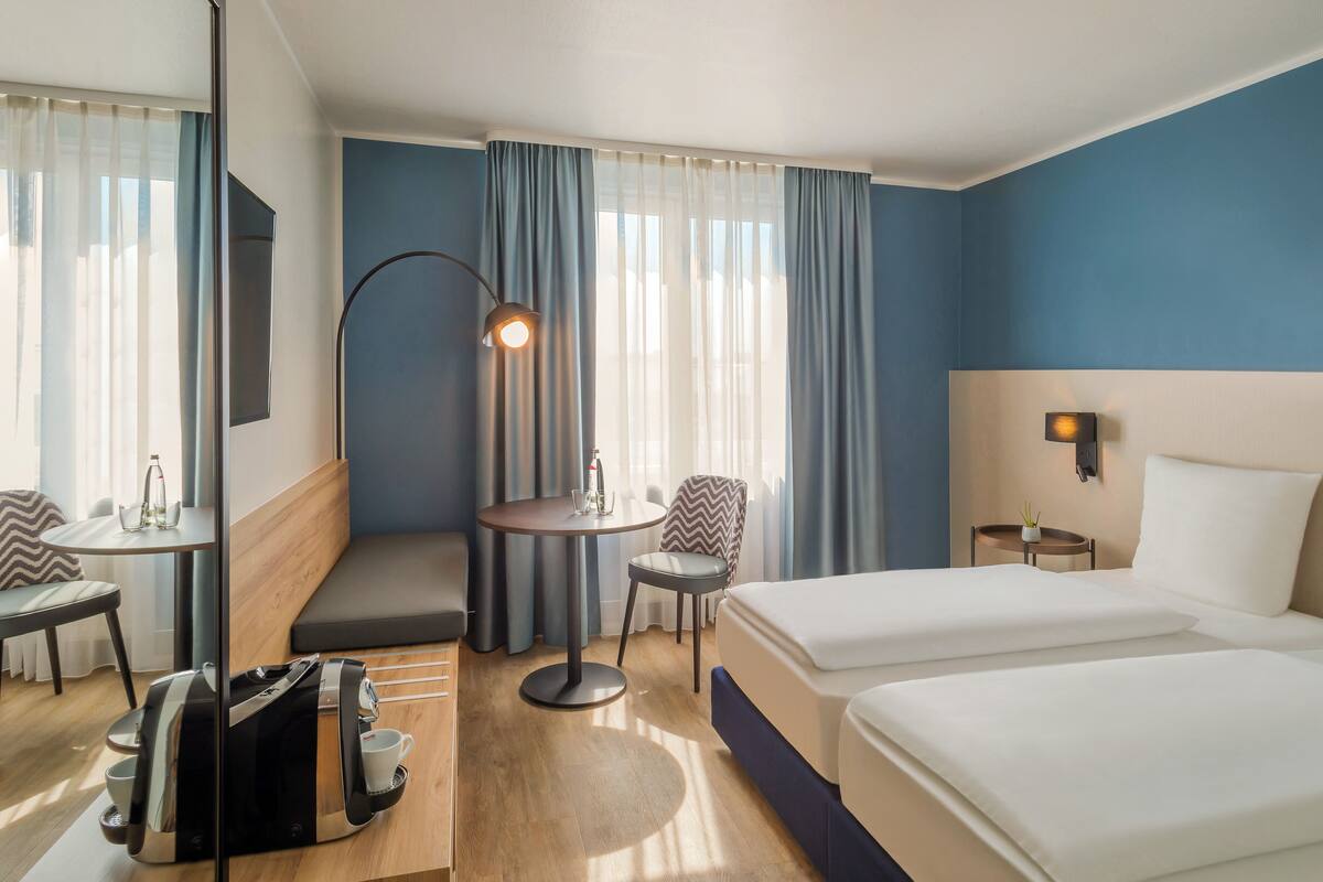 Habitación de hotel con cama, escritorio con silla, una mesita con lámpara y ventana con cortinas.