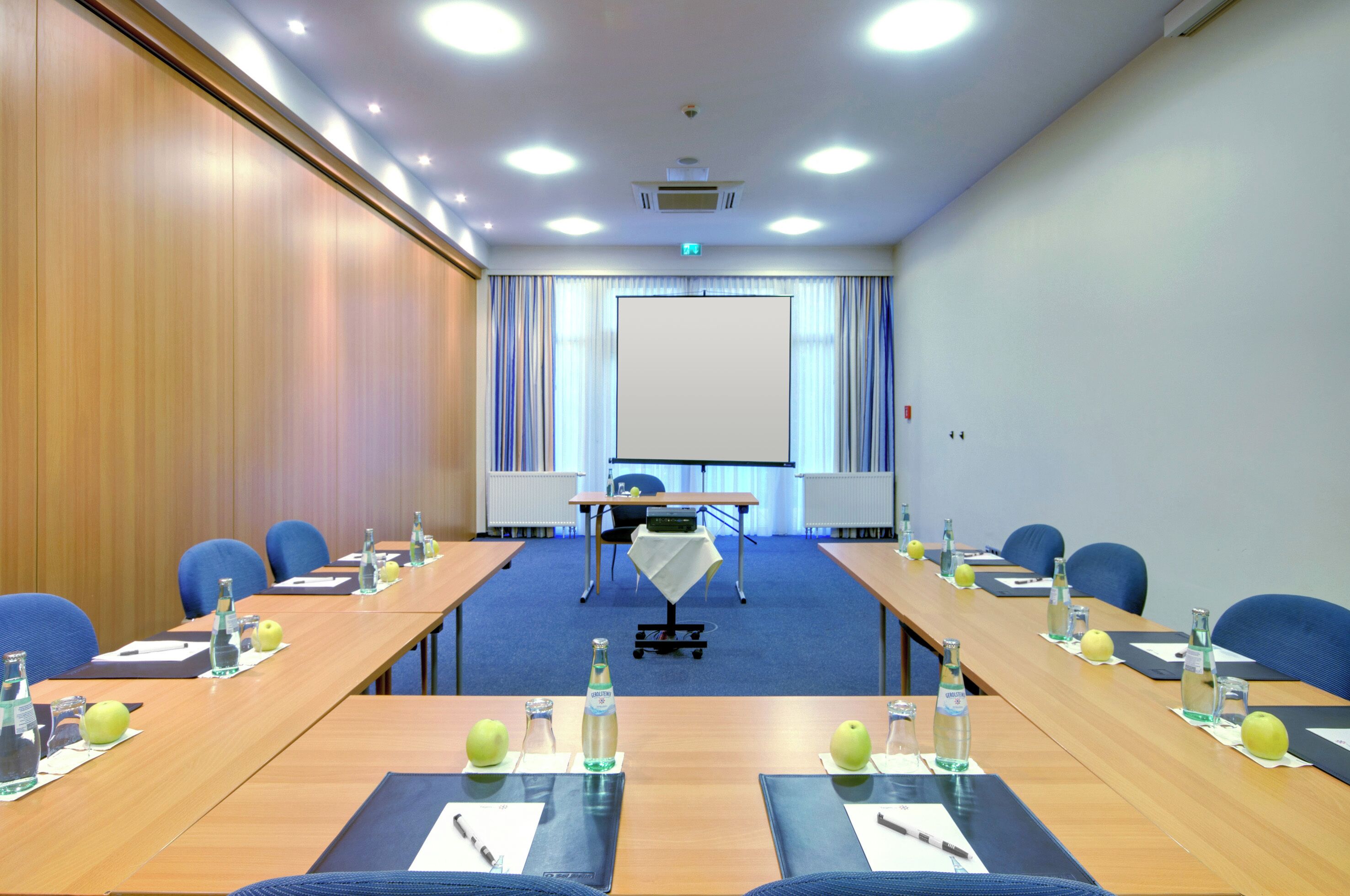 A conference room with a long table, chairs, a projector screen, and a table with water bottles and apples.