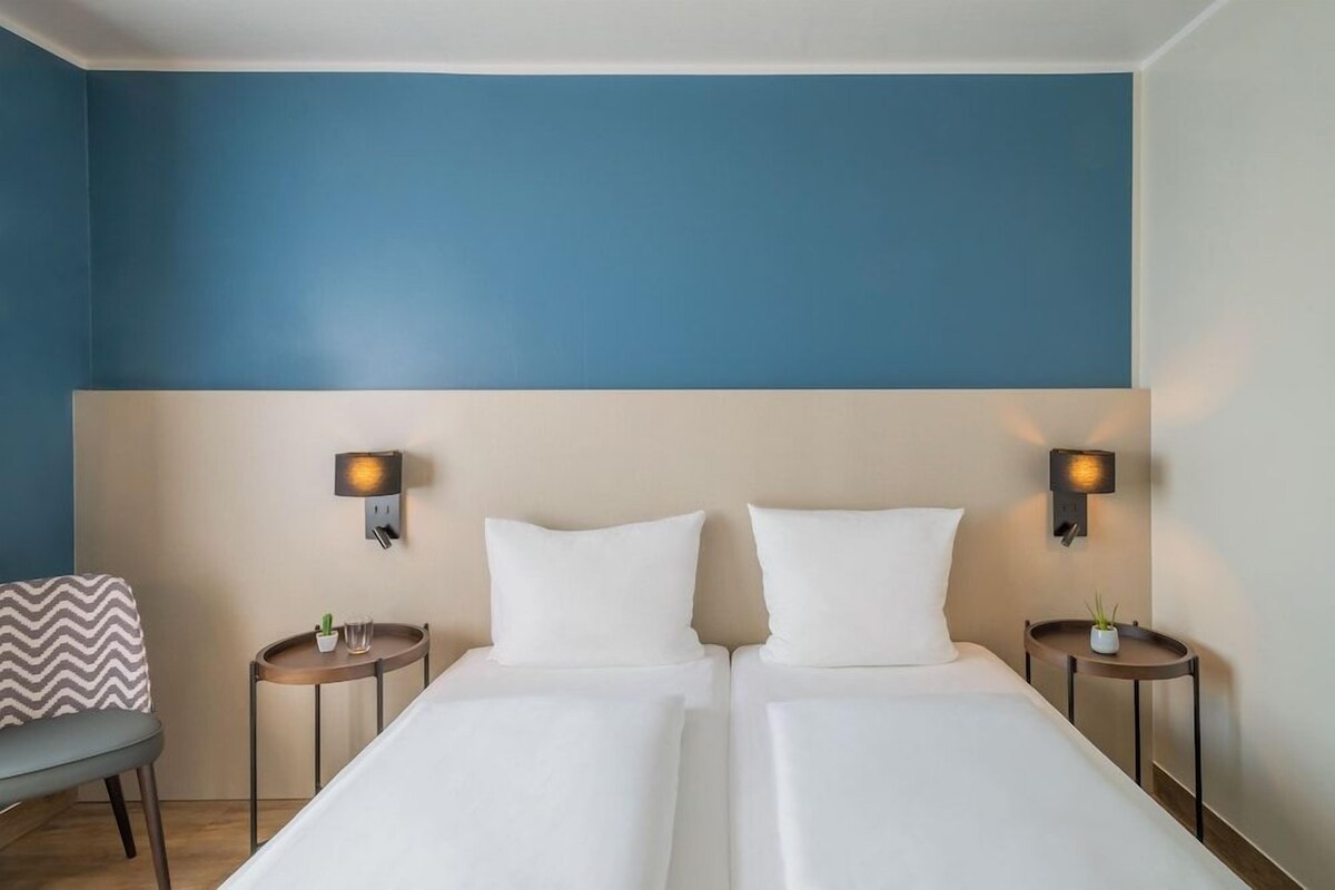 Habitación de hotel con dos camas, una silla y dos mesitas de noche con lámparas.