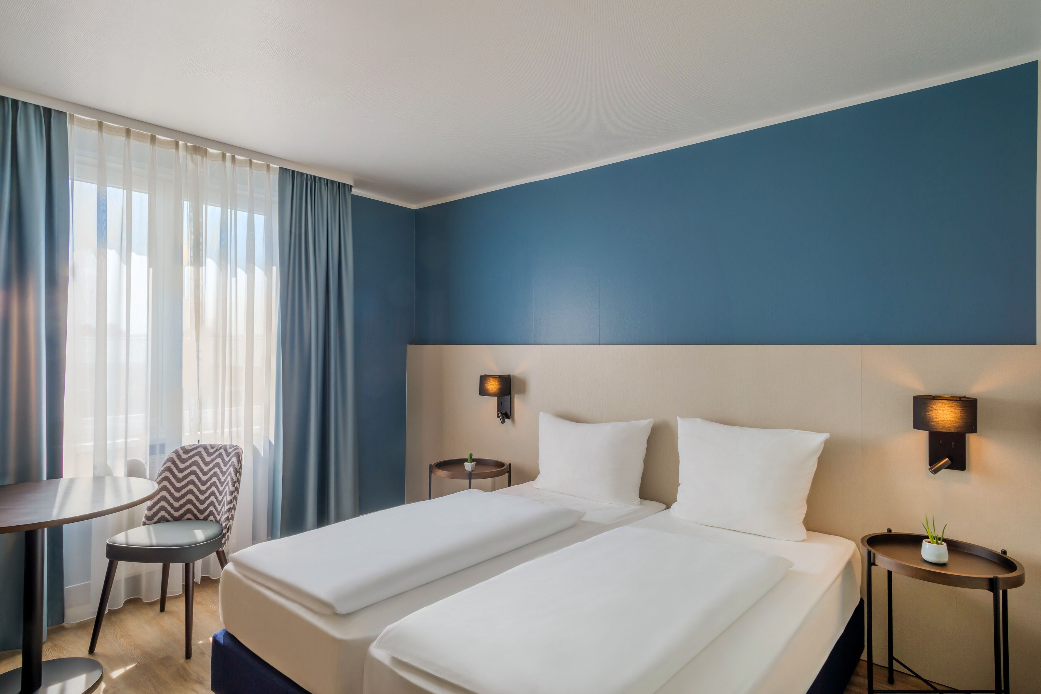 Ein Hotelzimmer mit zwei Betten, einem Schreibtisch, einem Stuhl und einem kleinen Tisch mit einer Pflanze.