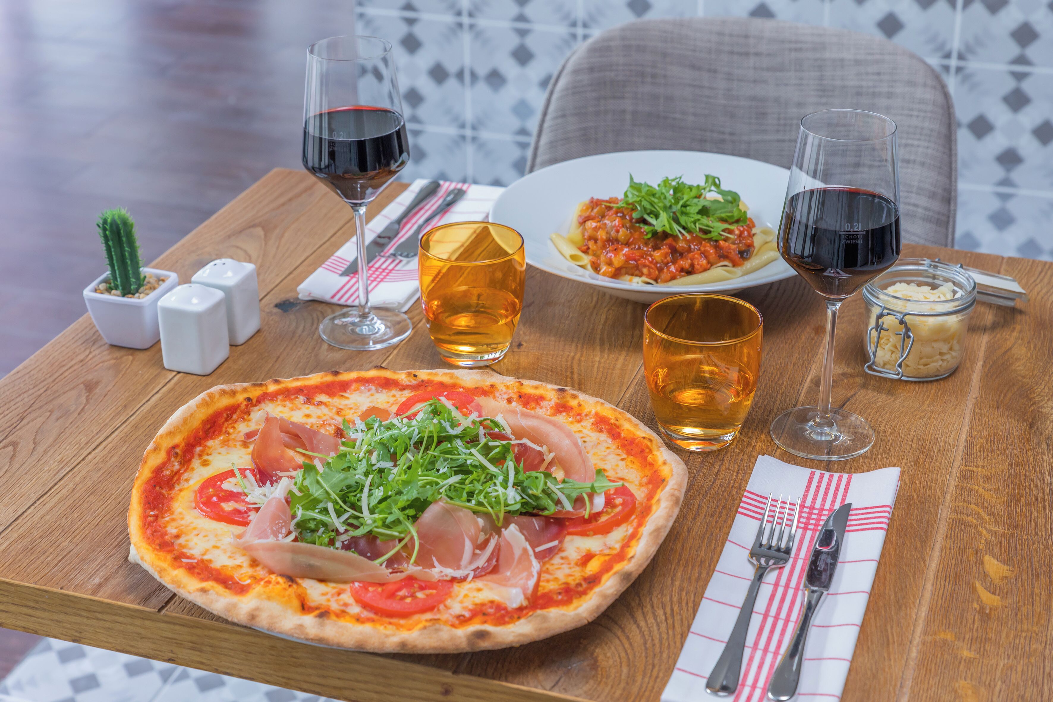 A wooden table with a pizza, a plate of pasta, and two glasses of red wine.