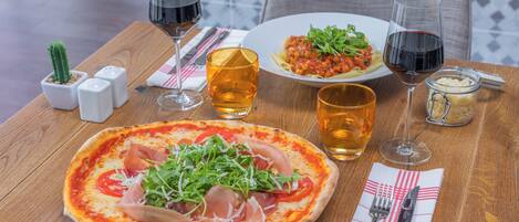 A wooden table with a pizza, a plate of pasta, and two glasses of red wine.