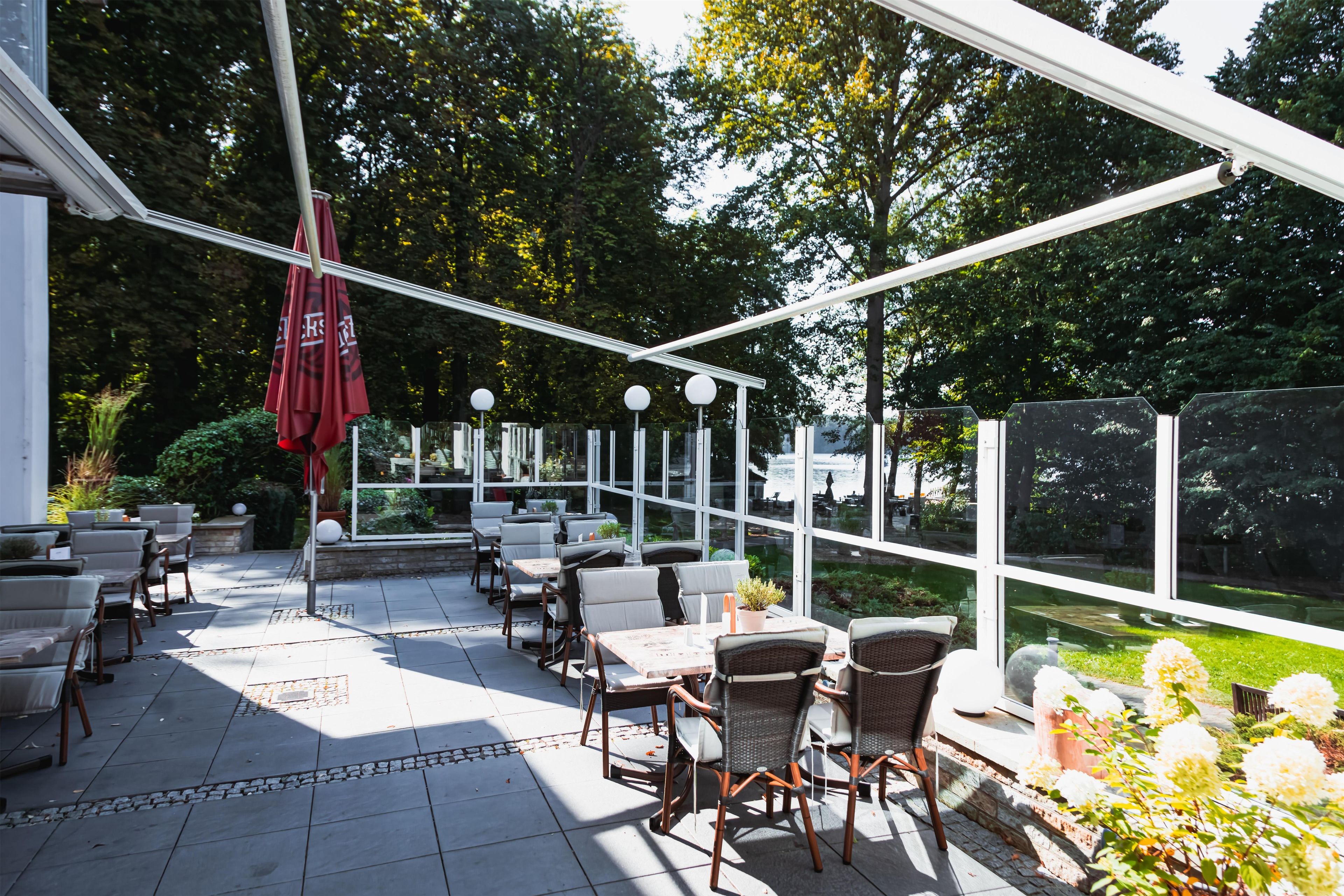 En udendørs terrasse med borde og stole, et glashus og udsigt til naturen.