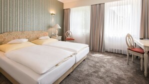 A hotel room with two beds, a desk, a chair, a lamp, and a view of the city through sheer curtains. - Seehotel Frankenhorst - BW Signature Collection (Schwerin)