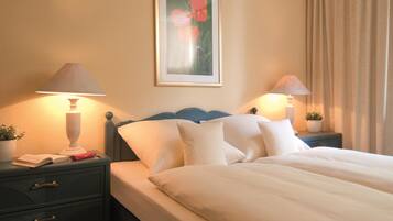 A hotel room with a bed, two bedside tables with lamps, a nightstand with a book and a red object, a framed picture on the wall, and a potted plant on the nightstand.