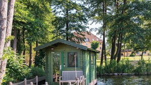 Une petite cabane en bois avec un banc sur un quai, au bord d’une étendue d’eau calme, entourée d’une végétation luxuriante et d’arbres.