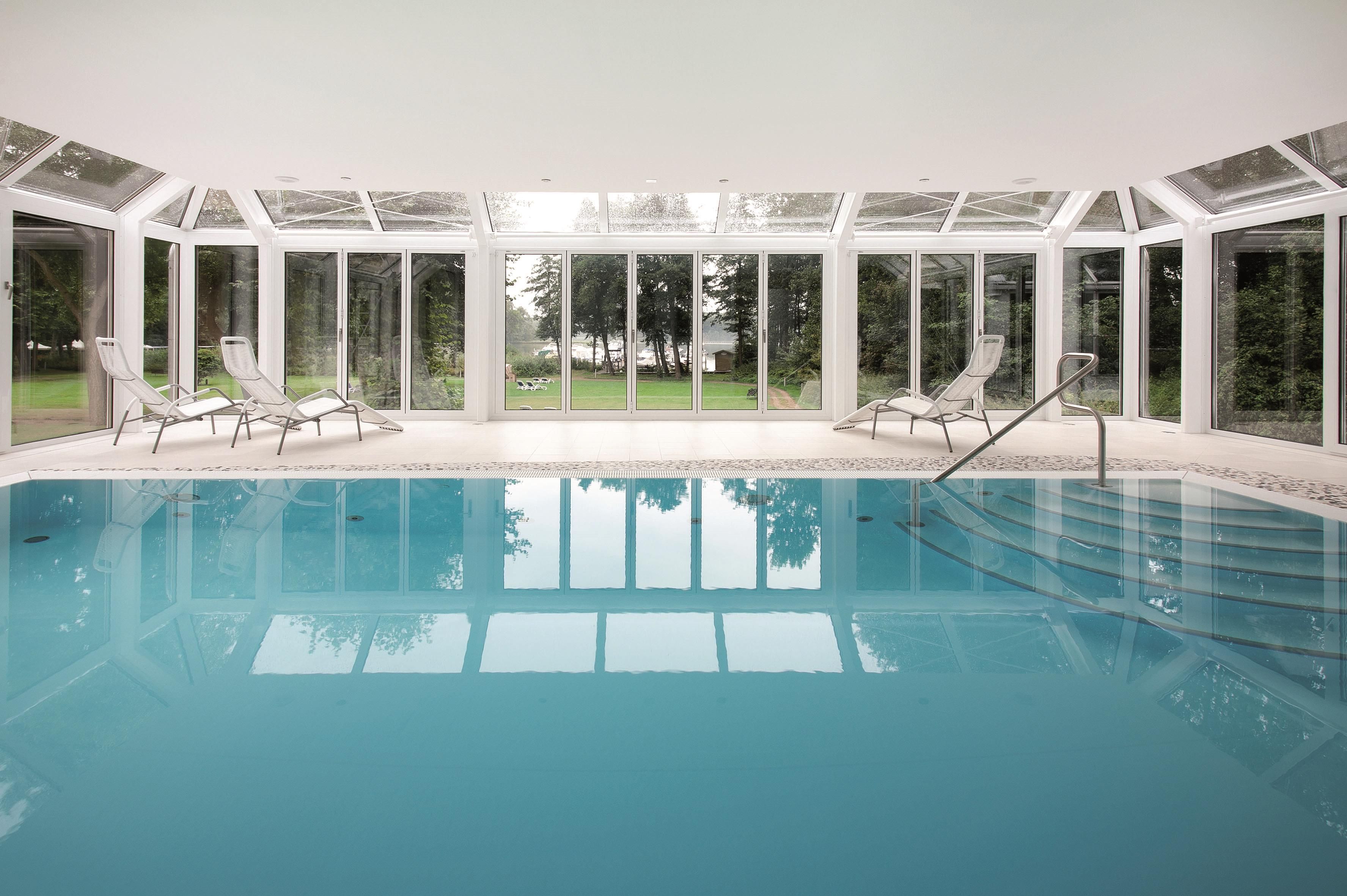 En swimmingpool med kristallklart blått vatten, omgiven av glasväggar och vita stolar.