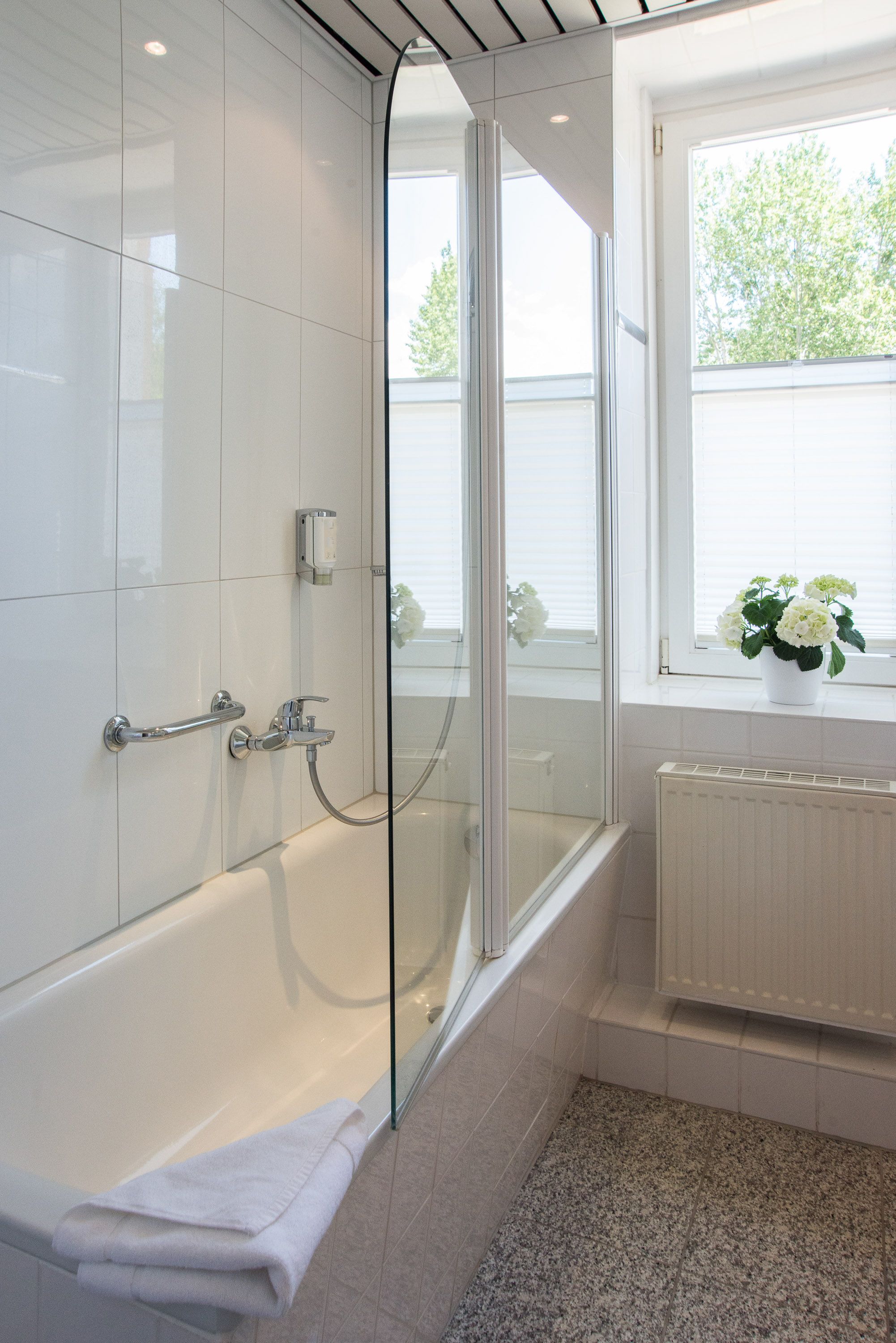 A modern bathroom with a glass-enclosed shower, a white bathtub, and a window with a view of greenery.
