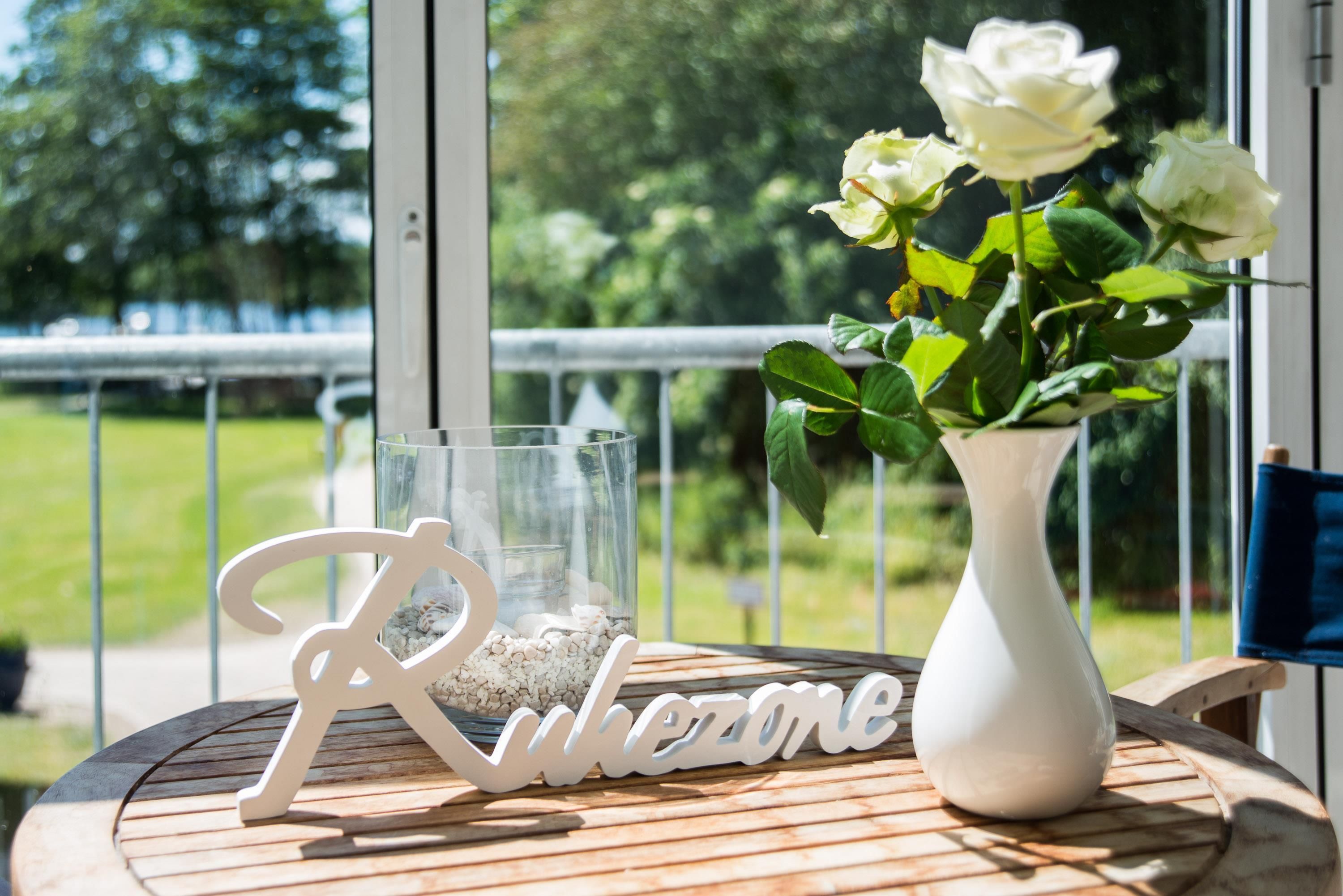 Una mesa con un jarrón blanco lleno de flores blancas, un vaso con piedras y un letrero de madera que dice 'Rulezone'.