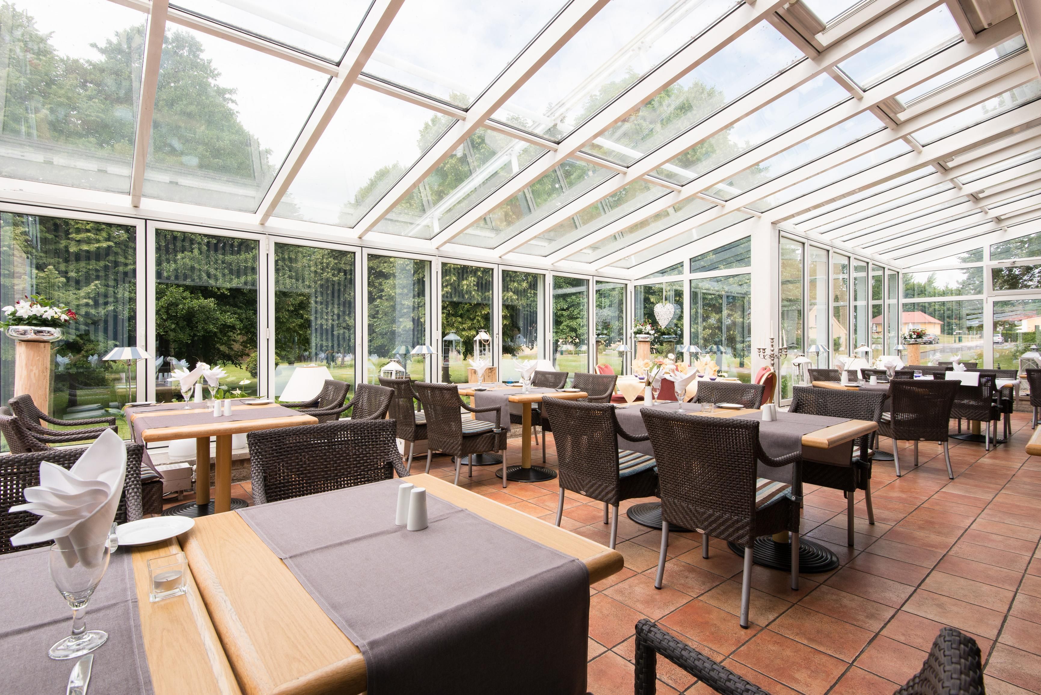 A conservatory with glass walls, furnished with tables and chairs, and a view of the outdoors.