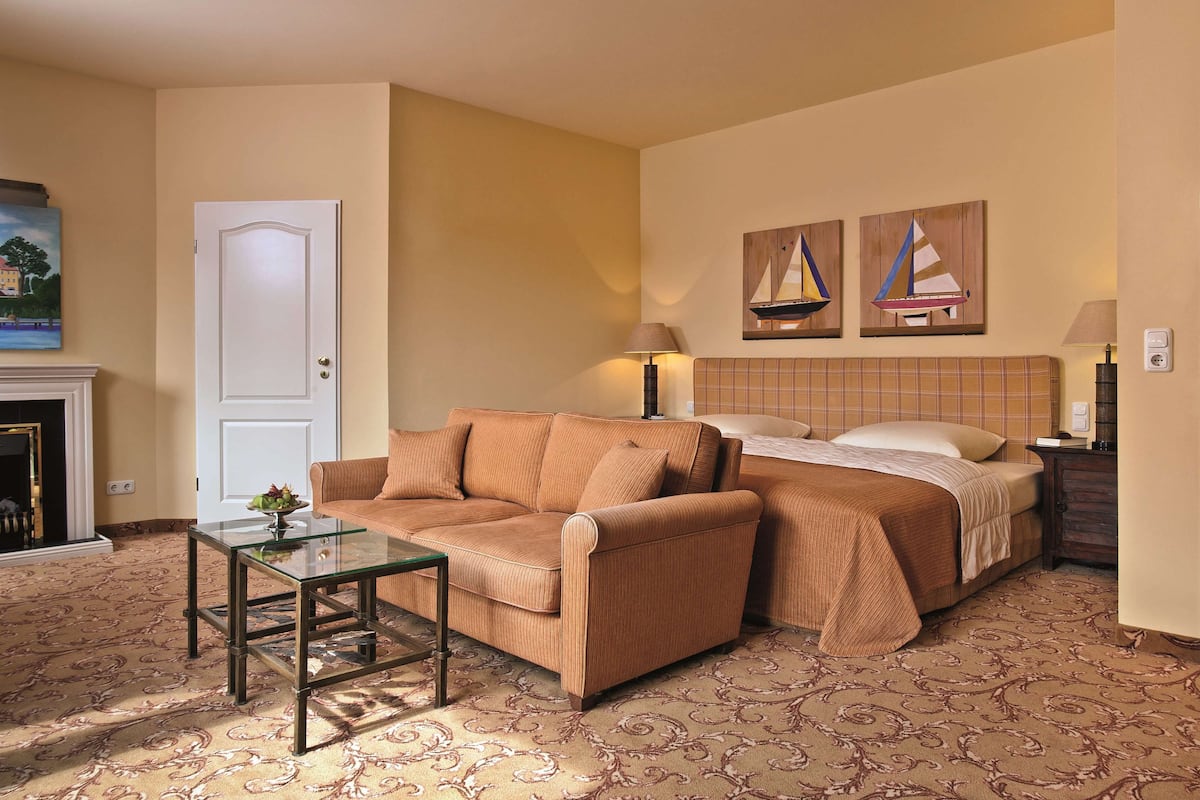 A hotel room with a sofa, a bed, a glass coffee table, and two paintings on the wall.