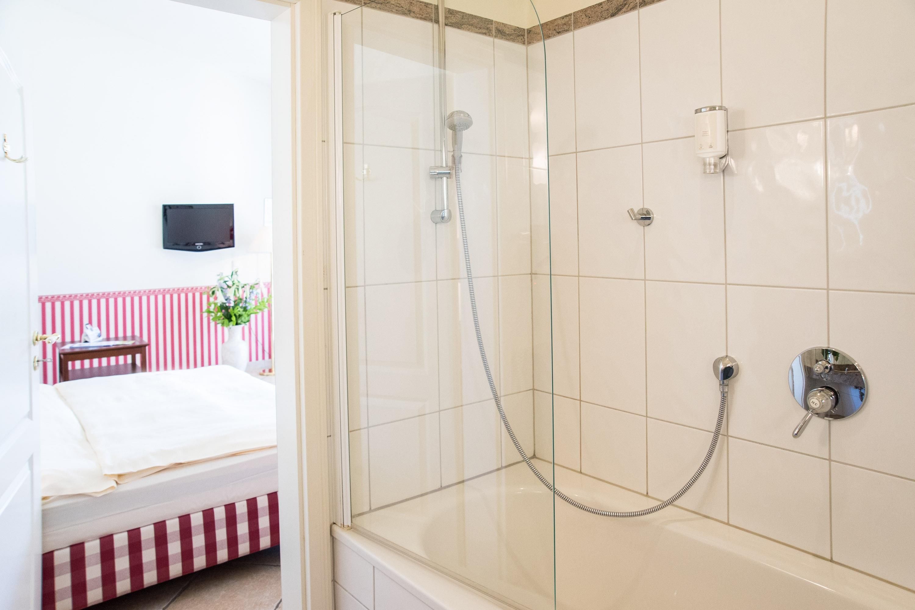 Un baño de hotel con ducha de vidrio, una cama con colcha a cuadros y un televisor montado en la pared.