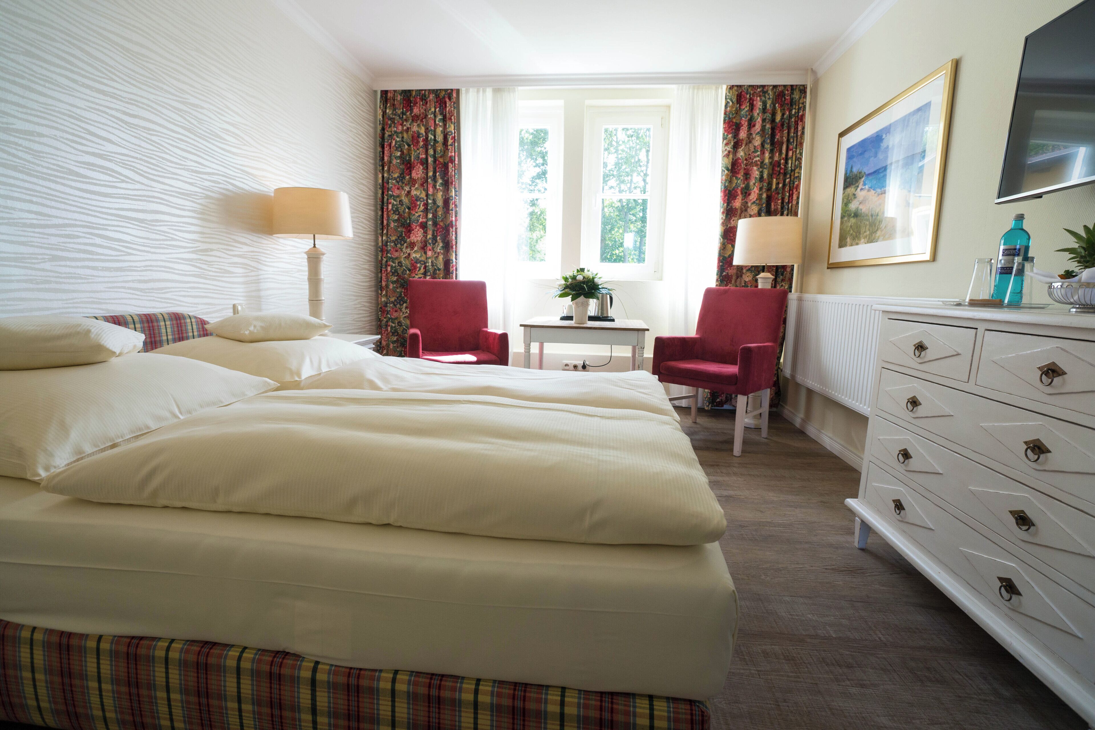 A hotel room with a large bed, two red chairs, a small table, a dresser, and a window with curtains.