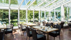 Une véranda aux murs de verre, aménagée avec des tables et des chaises, et entourée de verdure.