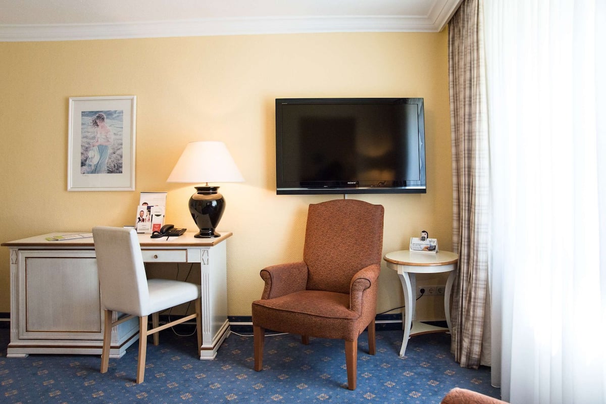 A hotel room with a desk, chair, lamp, television, and a small table with a remote control.