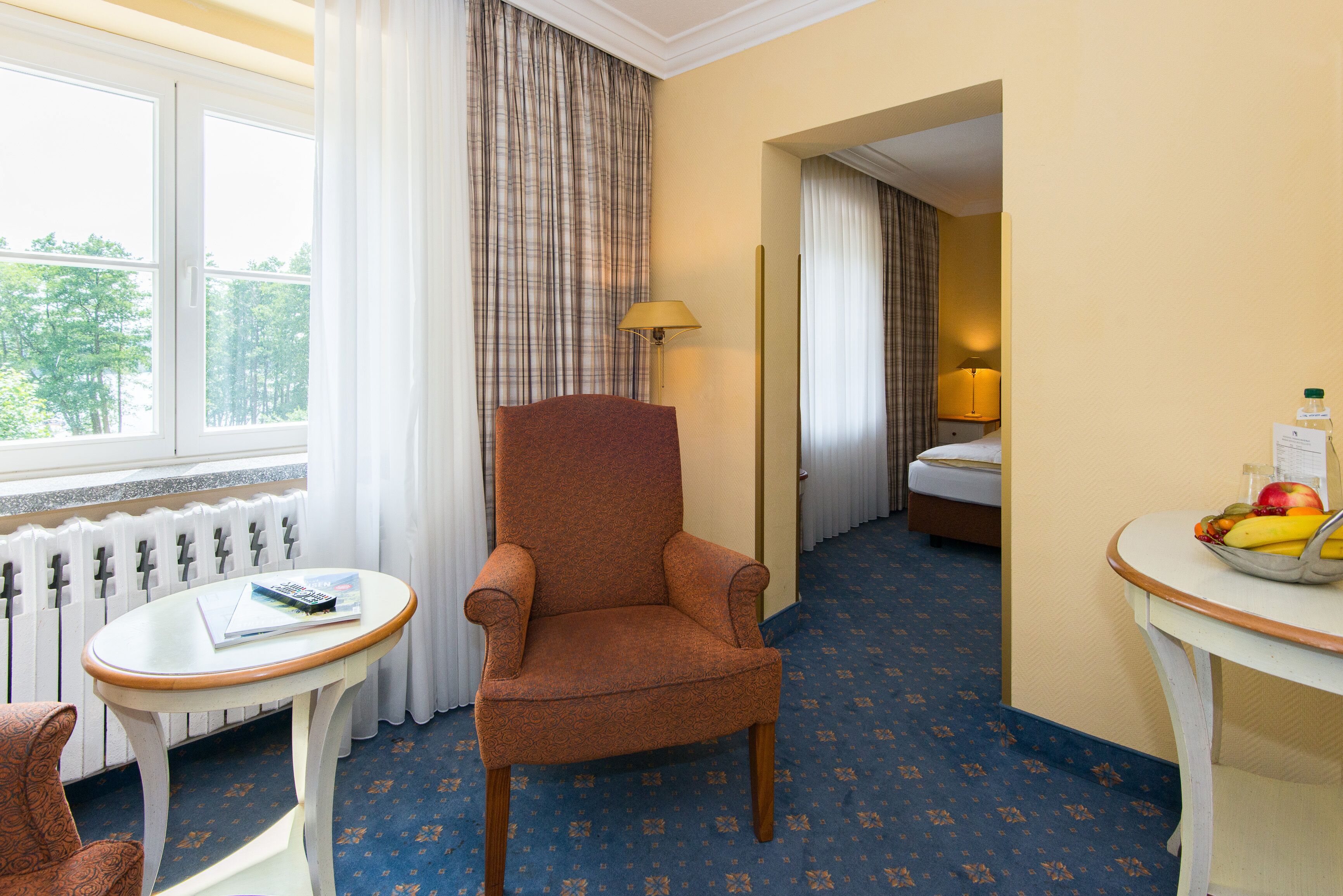 A hotel room with a large window, a plaid curtain, a brown armchair, a small round table with a book, a fruit bowl, and a bed with a white duvet.