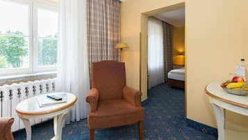 A hotel room with a large window, a plaid curtain, a brown armchair, a small round table with a book, a fruit bowl, and a bed with a white duvet.