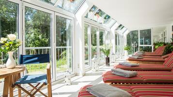 Una habitación soleada con ventanas amplias, una zona de estar con cojines a rayas y una pequeña mesa con un florero.