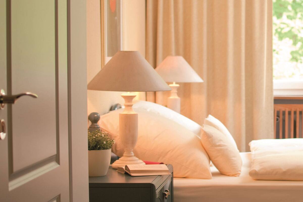 A neatly made bed with white linens and pillows, a bedside table with a lamp, a small potted plant, and a book.