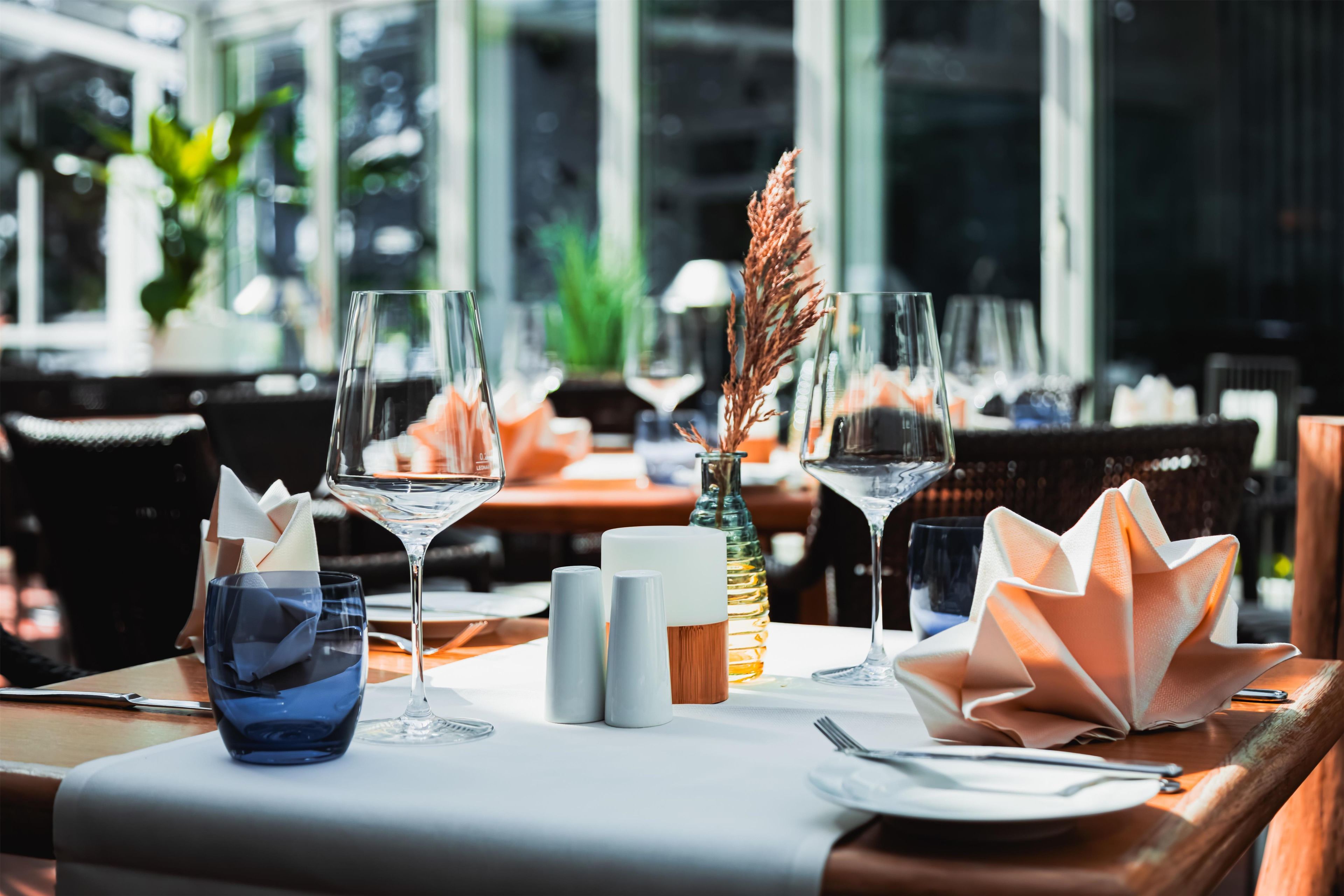 Ein mit Weingläsern, einer blauen Glasschale, Salz- und Pfefferschüttlern, einer Zierpflanze und einem gefalteten Servietten gedeckter Restauranttisch.