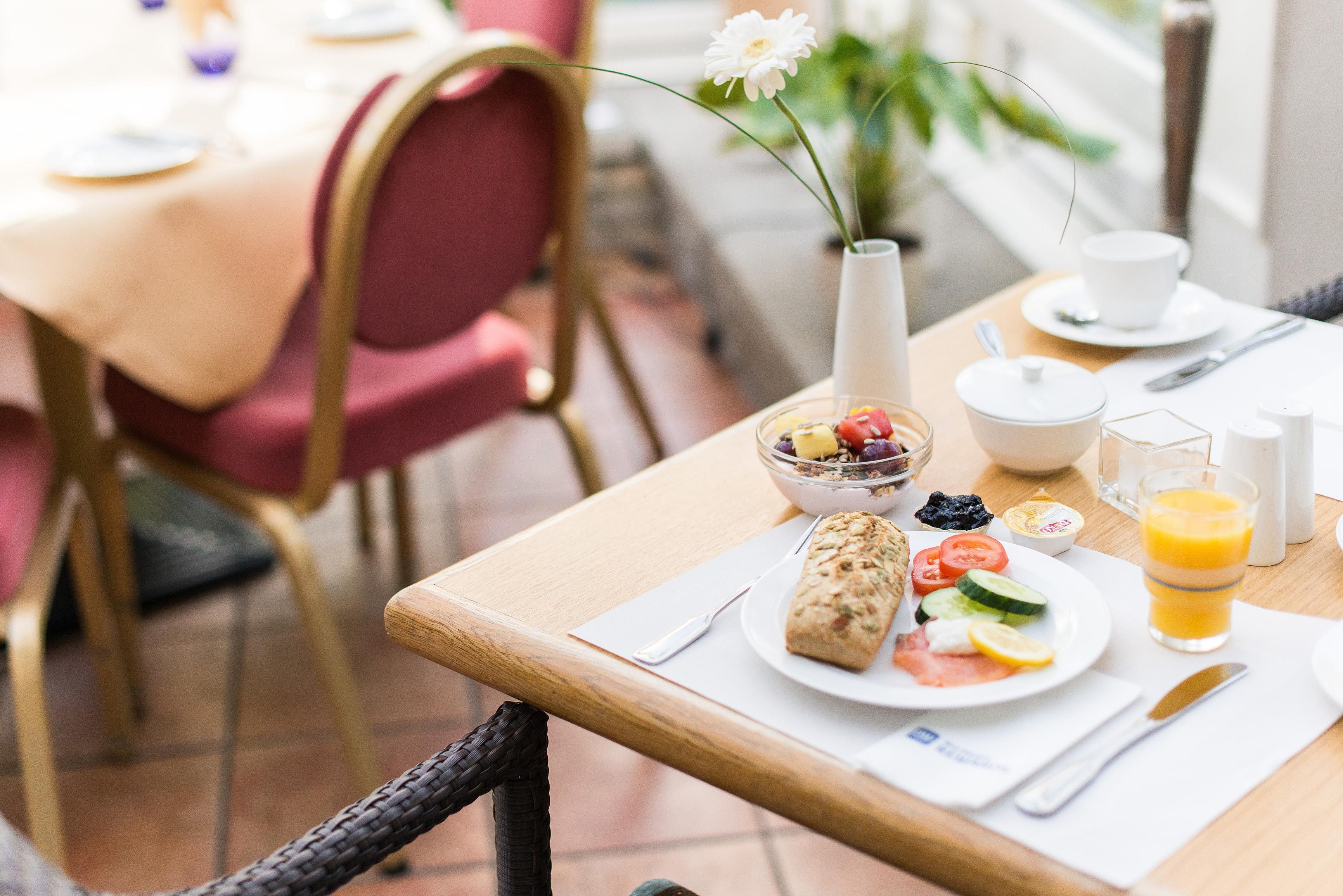 A breakfast table with a bowl of cereal, a glass of orange juice, a plate with sliced tomatoes and cucumbers, a wrapped pastry, and a small bowl of yogurt.