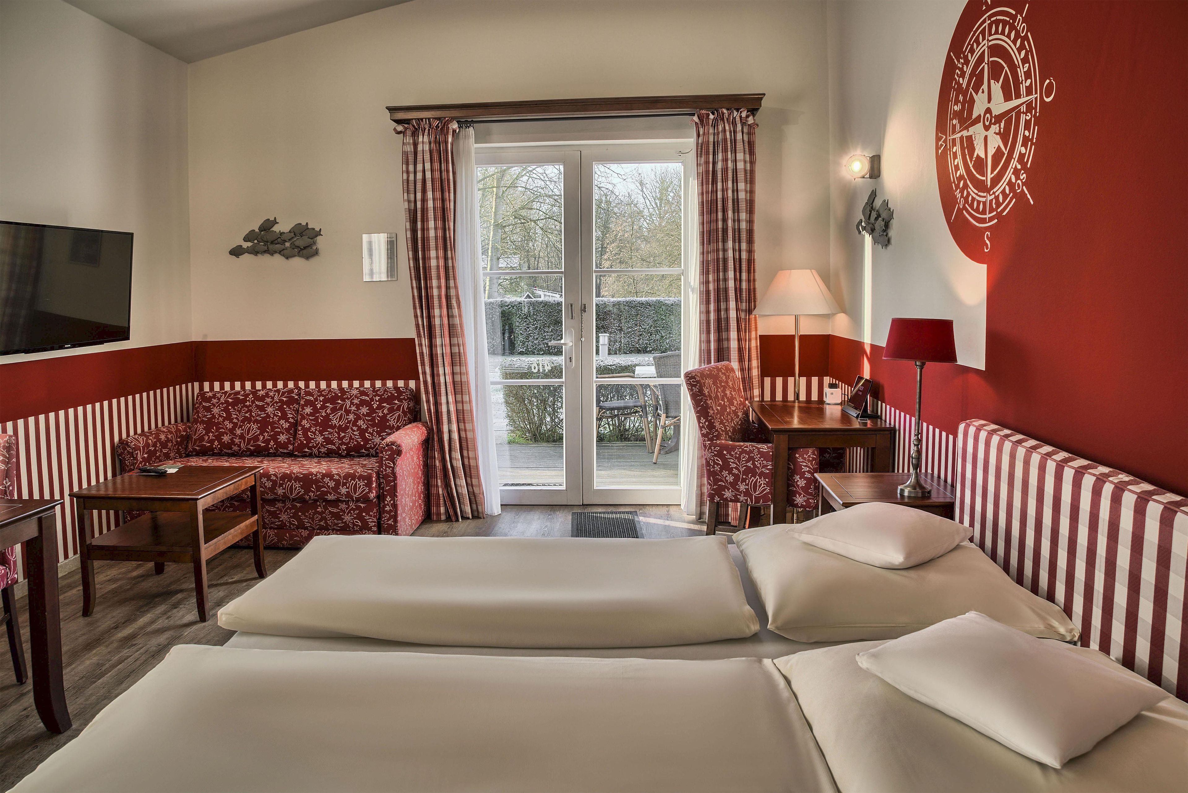 Habitación de hotel con cama, sofá, sillón, televisor y puerta corrediza de vidrio al exterior.