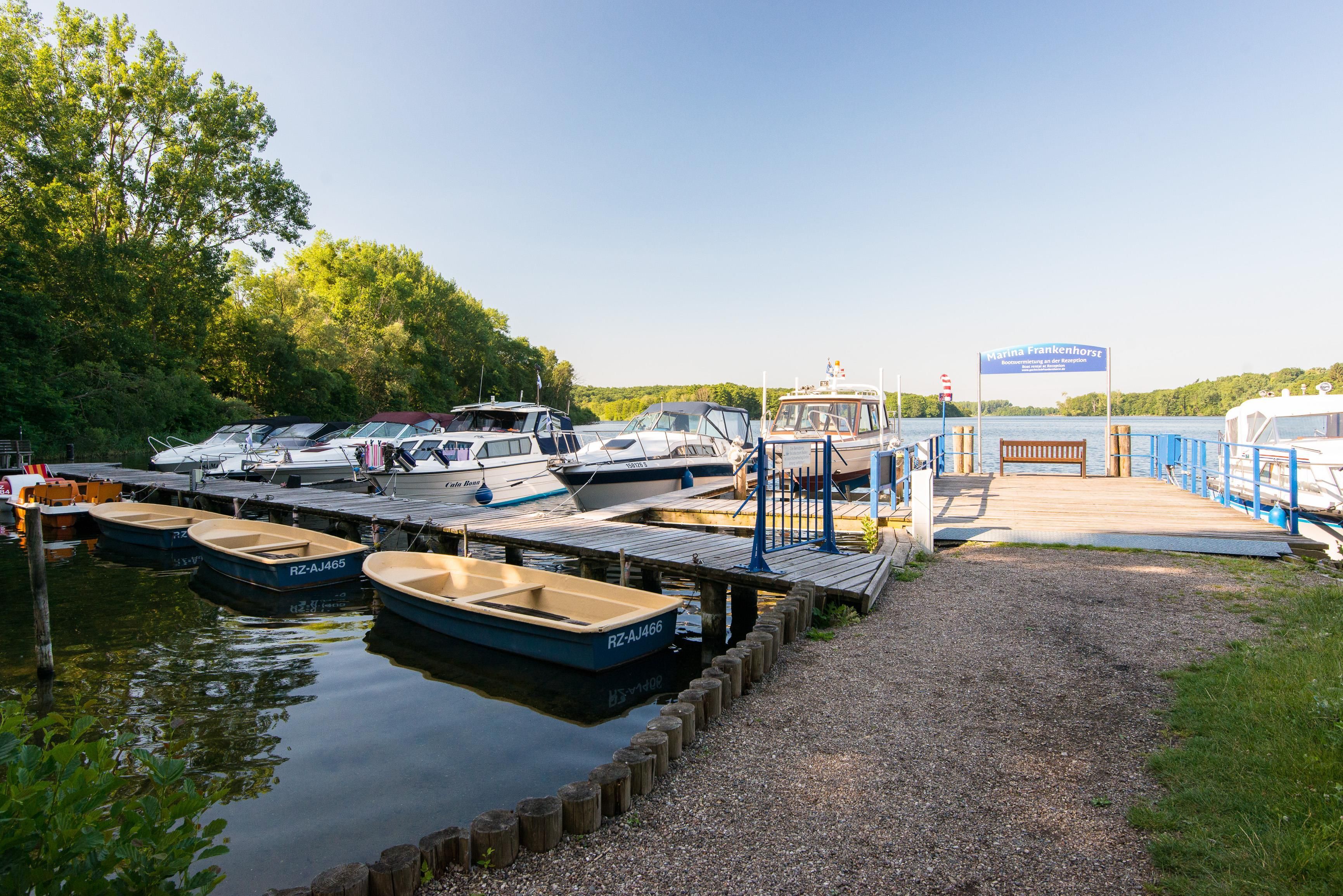 A marina with several boats docked, a wooden walkway leading to a reception area, and a sign indicating marina services.