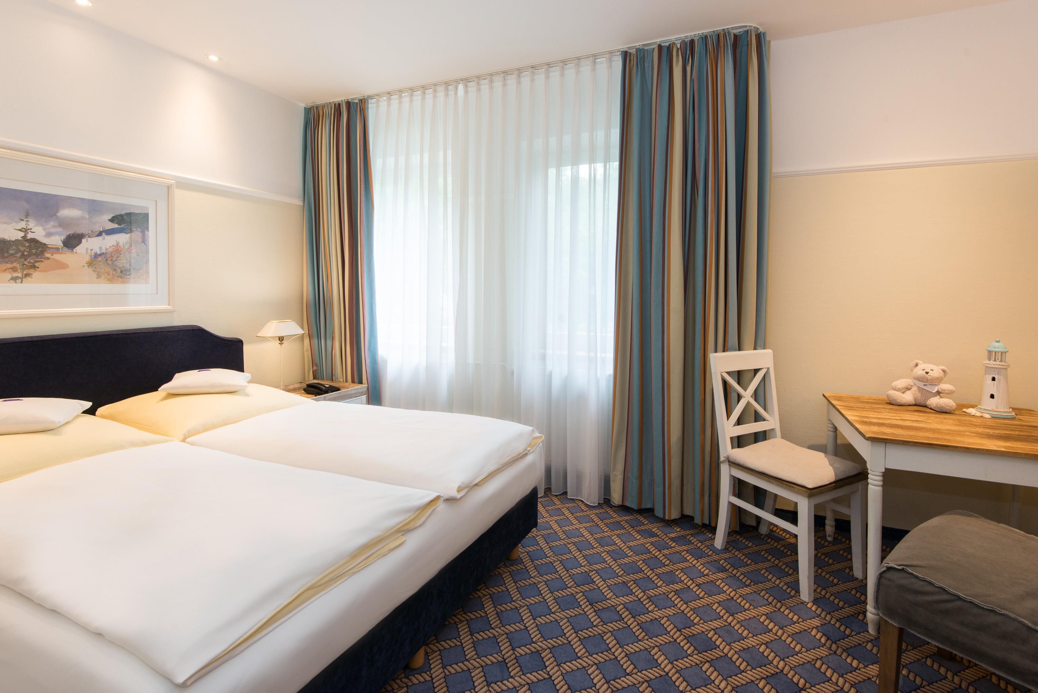 Quarto de hotel com duas camas, uma escrivaninha, uma cadeira, um abajur e um quadro na parede.