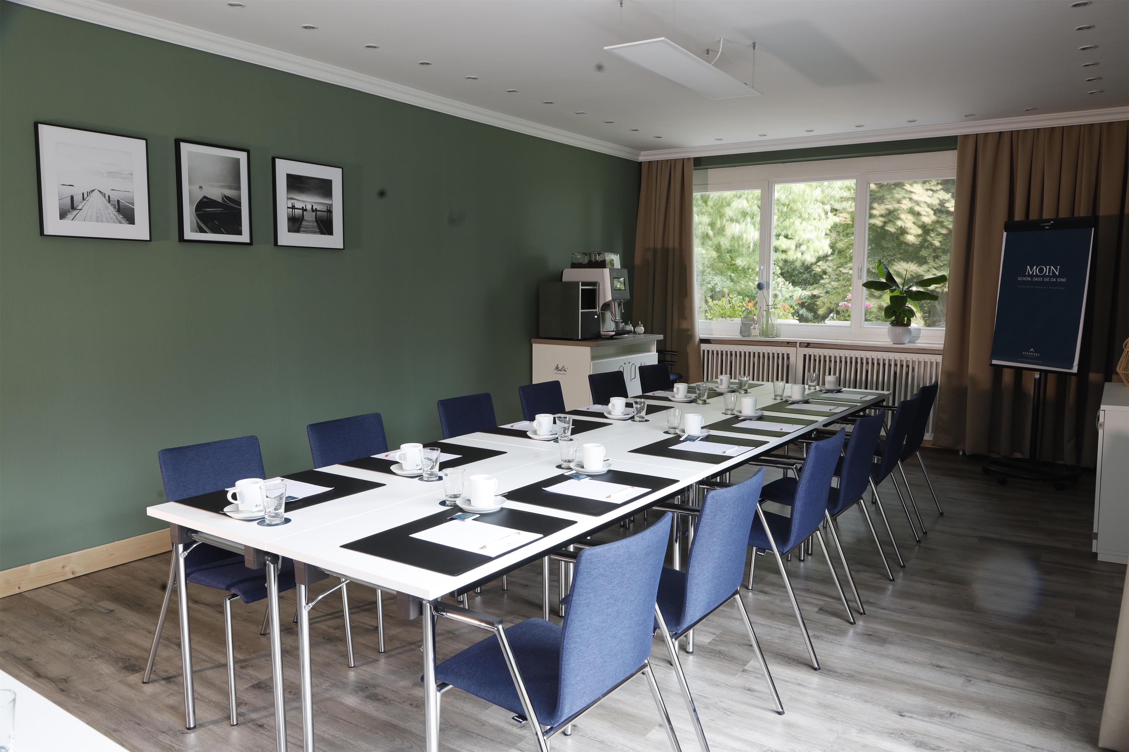 A conference room with a long table, chairs, a coffee station, and a view of greenery through the window.