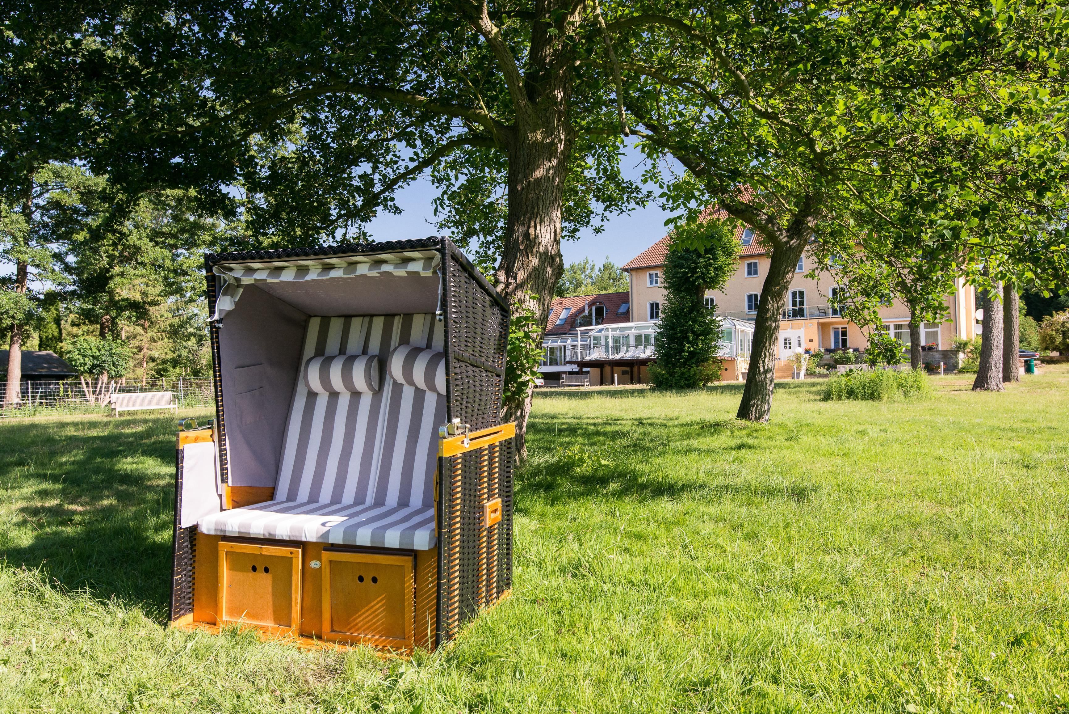 A small, enclosed outdoor seating area with a striped cushion and a wooden structure, surrounded by grass and trees.
